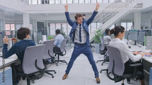 Young Cheerful Handsome Business Manager Wearing a Suit and Tie Dances in the Office. Diverse and Motivated Business People Work on Computers in Modern Open Office. photo
