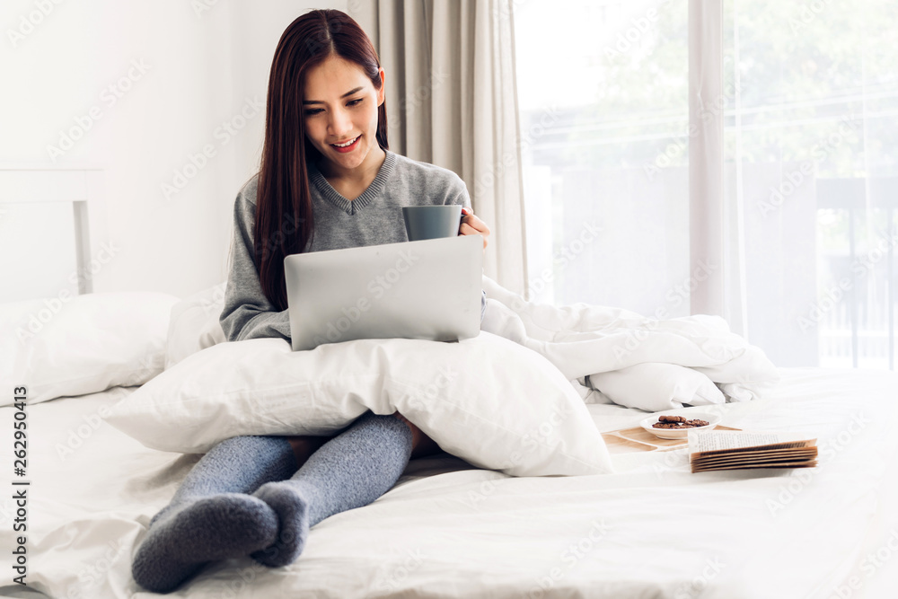 Young woman relaxing and drinking cup of hot coffee or tea using laptop computer on a cold winter day in the bedroom.woman checking social apps and working.Communication and technology concept Young woman relaxing and drinking cup of hot coffee or tea using laptop computer on a cold winter day in the bedroom.woman checking social apps and working.Communication and technology concept