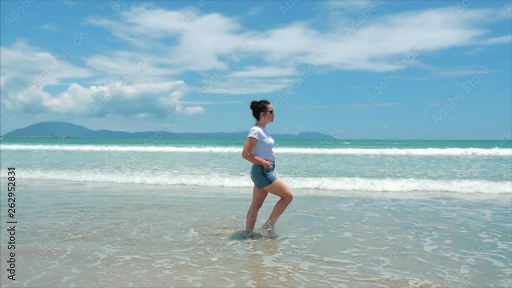 European Beautiful Brunette in Black Glasses, White-shirt and Blue Shorts Goes Under the Hot Summer Sun Along the Tropical Exotic Coast of Vietnam, Carefree Girl Walking on the Sand Right to the Ocean European Beautiful Brunette in Black Glasses, White-shirt and Blue Shorts Goes Under the Hot Summer Sun Along the Tropical Exotic Coast of Vietnam, Carefree Girl Walking on the Sand Right to the Ocean