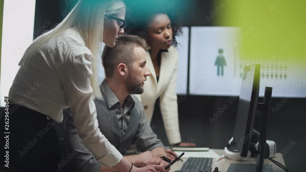 international teamwork in modern office, man and two women managers look at the screen with demographic infographics in monitor, camera movement through glass with sticky notes. international teamwork in modern office, man and two women managers look at the screen with demographic infographics in monitor, camera movement through glass with sticky notes.