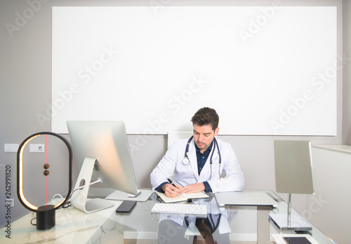Doctor sitting at the desk of his clinic office writing on his paper agenda. White blank poster with copy space in the background.