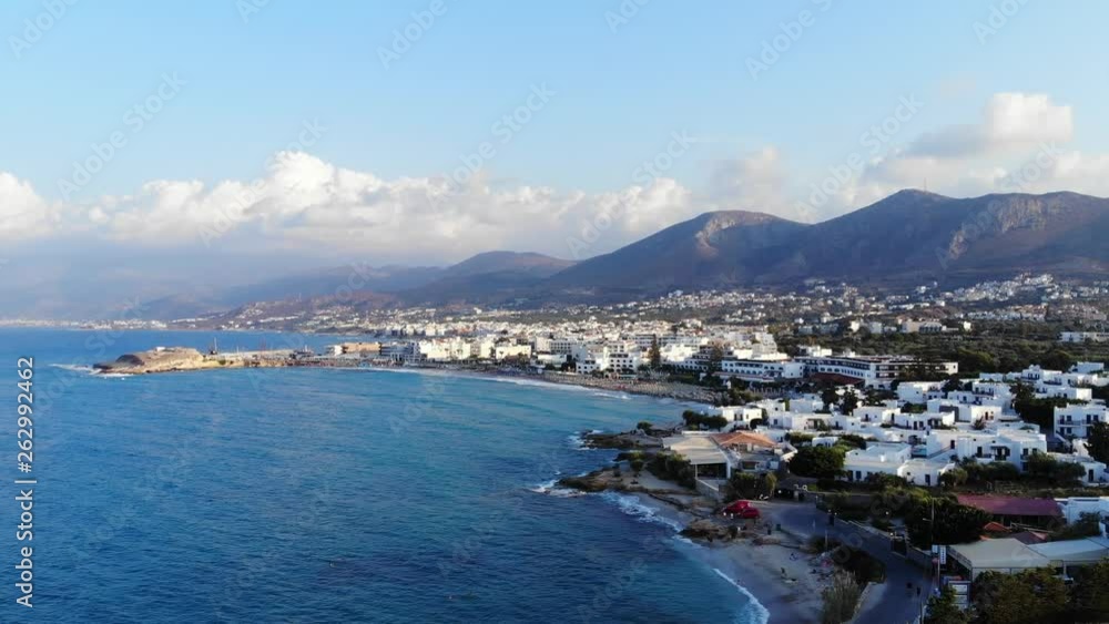 Aerial view of typical touristic town at North shore of Crete Island, camera turn right. Azure Aegean sea, low rise buildings at water front, mountain range seen on background Aerial view of typical touristic town at North shore of Crete Island, camera turn right. Azure Aegean sea, low rise buildings at water front, mountain range seen on background