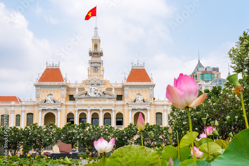 Saigon City Hall with pink lotus flowers (blurred) and blooming plumeria trees in the foreground. One of the top tourist attractions of the city of Ho Chi Minh City, Vietnam.