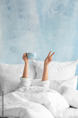 Young woman with cup of hot beverage showing victory gesture in bed