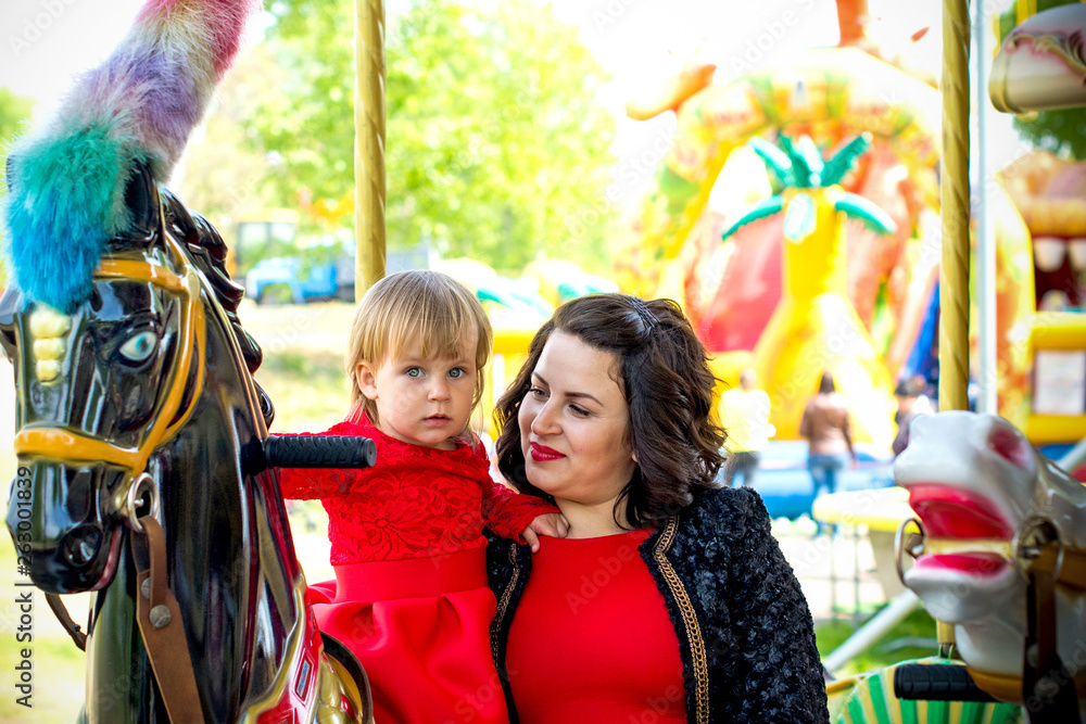 A stylish mother in a red dress and a black jacket and a little daughter in a leather jacket for a walk in the city in an amusement park. The family is having fun and experiencing life