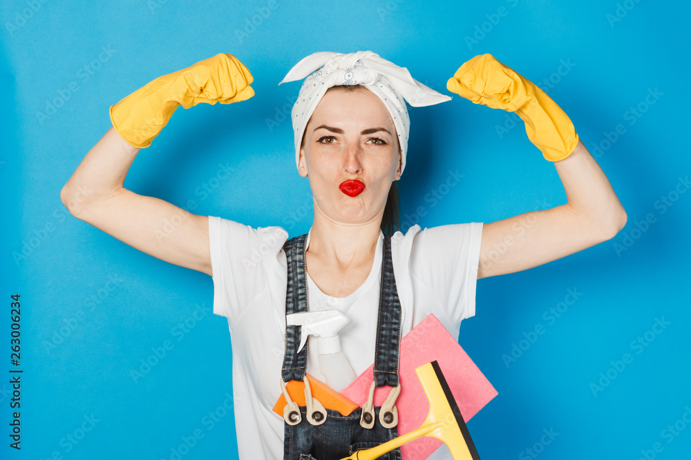 A young girl with a scarf on her head, yellow rubber gloves shows muscle biceps on her arm against a blue background. The concept of cleaning and cleaning service, high quality.