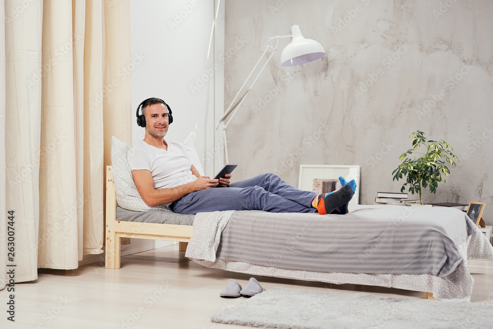 Caucasian middle aged man dressed casual relaxing on the bed in bedroom, listening music and using tablet.