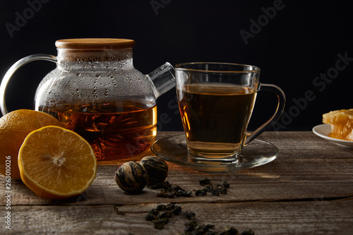 transparent teapot and cup with traditional blooming tea on wooden table with lemons isolated on black