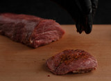 Hand in black glove sprinkling spice on beef steak on a wooden board with a black background
