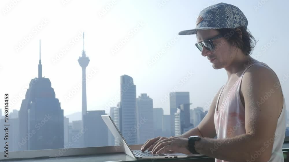 Freelancer using laptop on modern city background during summer vacation. Young man working online on skyscraper. Concept of: freelance, blogging, online education. Freelancer using laptop on modern city background during summer vacation. Young man working online on skyscraper. Concept of: freelance, blogging, online education.