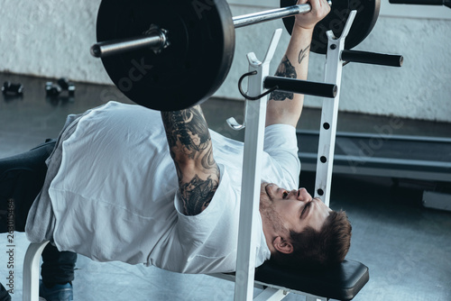 Overweight tattooed man in white t-shirt exercising with barbell at gym photo