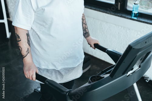 partial view of overweight tattooed man running on treadmill at sports center