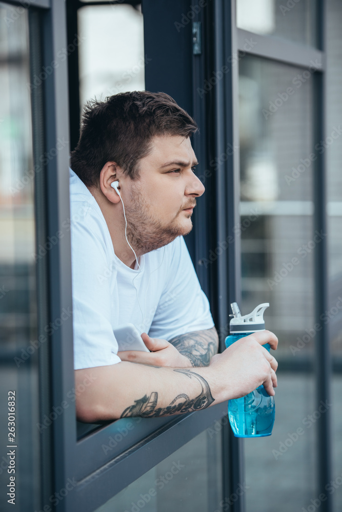 overweight man with earphones and smartphone holding sport bottle and looking out through window at gym overweight man with earphones and smartphone holding sport bottle and looking out through window at gym