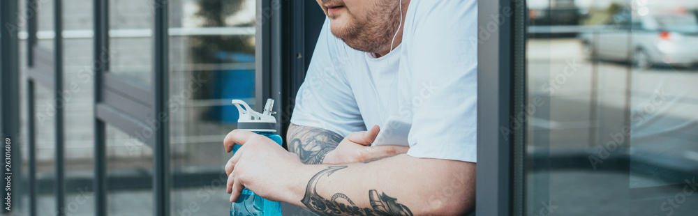 panoramic shot of overweight man with earphones and smartphone holding sport bottle and looking out through window at gym