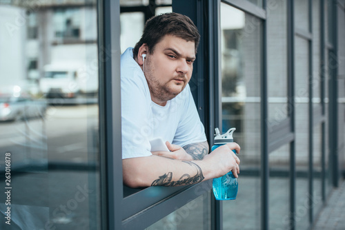 obese man with earphones and smartphone holding sport bottle and looking out through window at gym