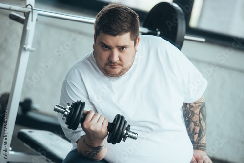 Overweight tattooed man Looking At Camera and training with dumbbell at sports center