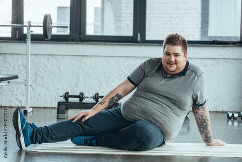 overweight tattooed man looking at camera, smiling and sitting on fitness mat at sports center