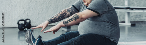 panoramic shot of overweight tattooed man sitting and stretching at gym