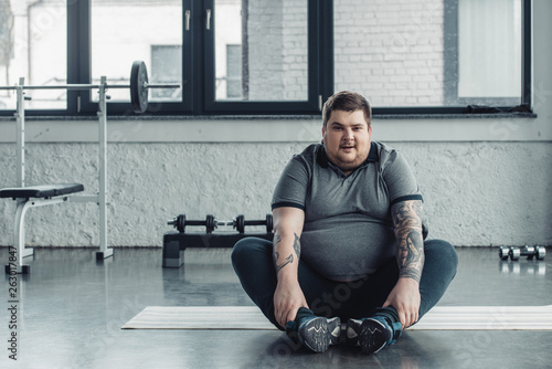 overweight tattooed man sitting and Looking At Camera during stretching exercise at gym photo