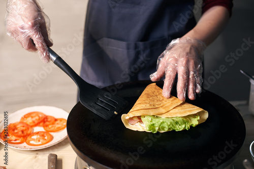 Chef in gloves making crepe on hot round portable cooktop. Skillful hands with spatula in final stage of cooking crepe stuffed with vegetables. Tomato slices on background. Healthy food. Close up view