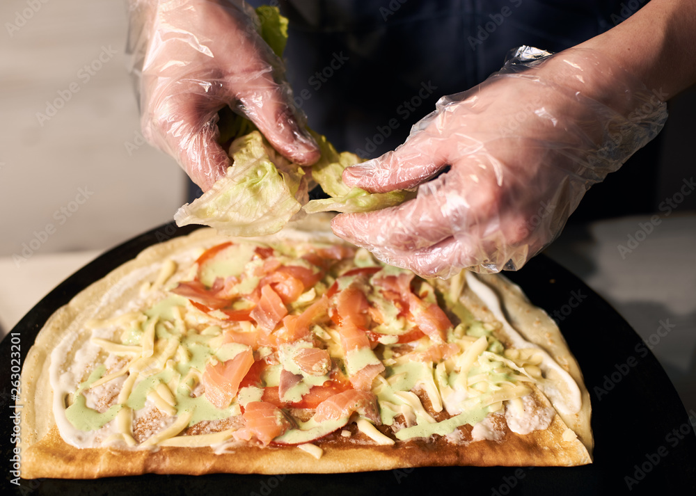 Chef's cropped hands in disposable gloves tearing green leaves of lettuce on round thin baked base for pizza cooking. Chopped pieces of fish, tomato, grated cheese on crepe on black tray. Close up.