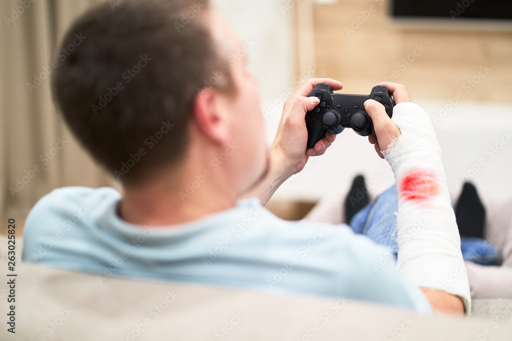 Man with broken arm in plaster cast with red blood play and holding controller in front of TV. Game addiction concept. Man with broken arm in plaster cast with red blood play and holding controller in front of TV. Game addiction concept.