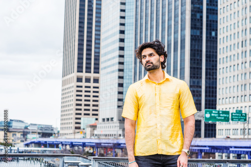 Raining day - grainy, wet feel. Young East Indian American Man with beard traveling in New York City, wearing yellow short sleeve suit, standing in front of modern high buildings, looking around..
