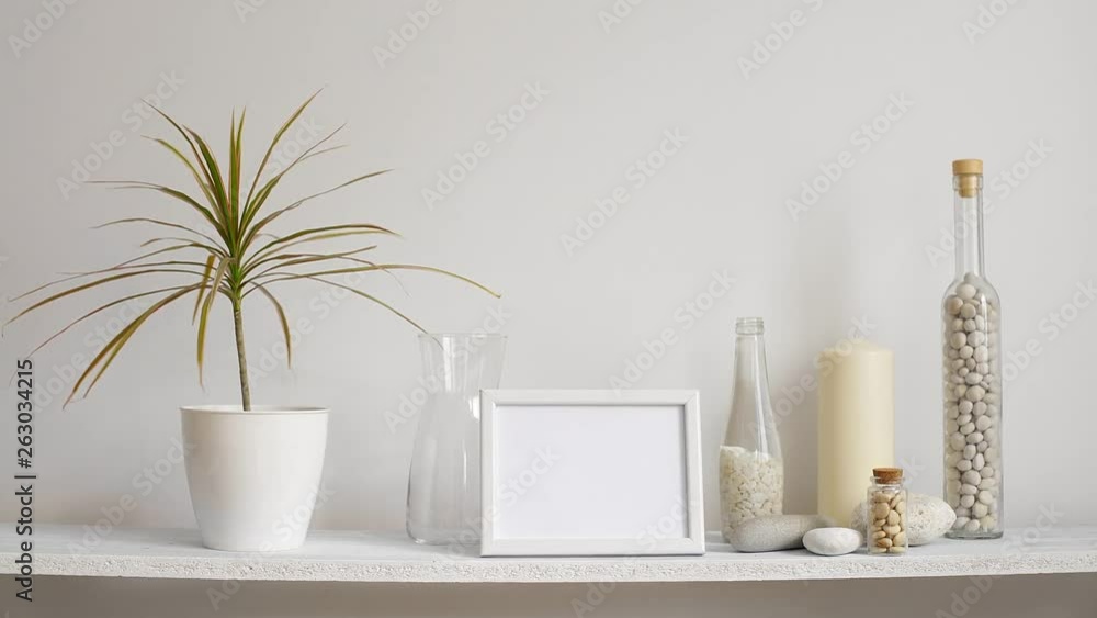 Modern room decoration with picture frame mockup. Shelf against white wall with decorative candle, glass and rocks. Hand putting down potted dracaena plant. Modern room decoration with picture frame mockup. Shelf against white wall with decorative candle, glass and rocks. Hand putting down potted dracaena plant.