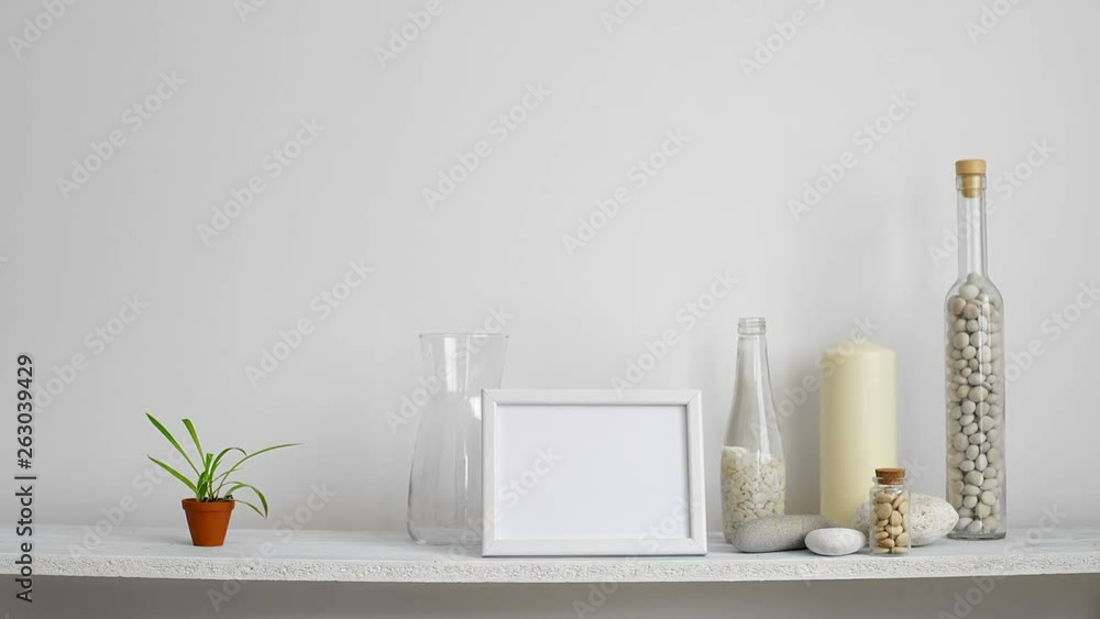 Modern room decoration with picture frame mockup. Shelf against white wall with decorative candle, glass and rocks. Hand putting down potted spider plant. Modern room decoration with picture frame mockup. Shelf against white wall with decorative candle, glass and rocks. Hand putting down potted spider plant.