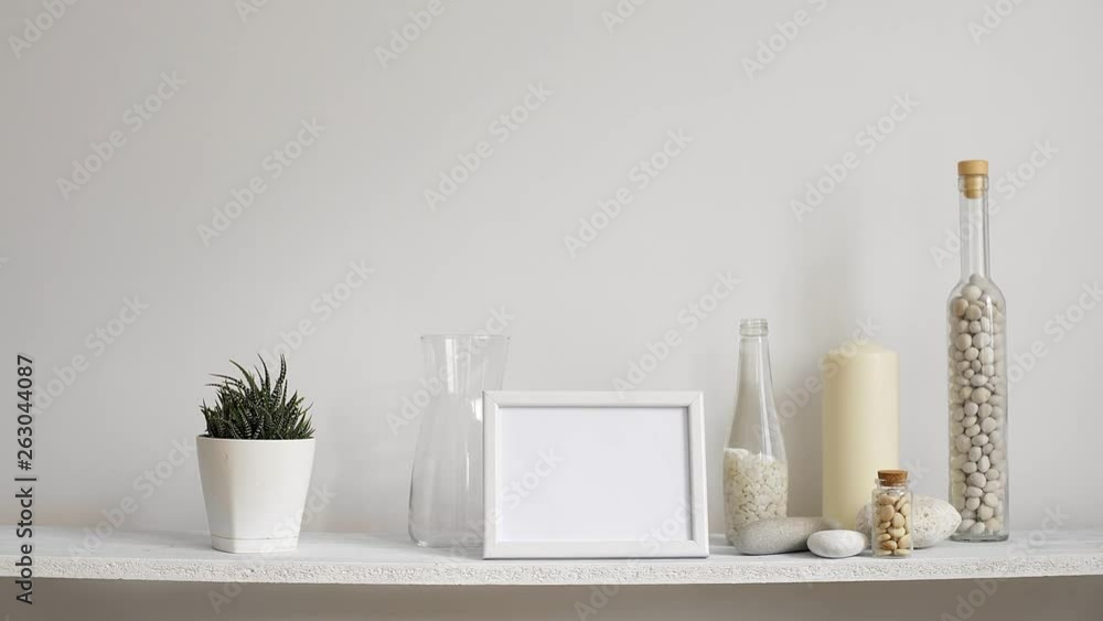 Modern room decoration with picture frame mockup. Shelf against white wall with decorative candle, glass and rocks. Hand putting down potted succulent plant. Modern room decoration with picture frame mockup. Shelf against white wall with decorative candle, glass and rocks. Hand putting down potted succulent plant.