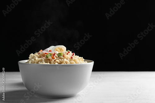 Bowl of hot noodles with egg and vegetables on table against black background. Space for text