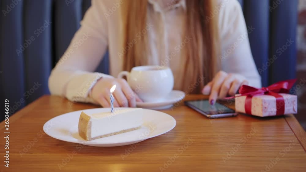 Close-up of waitress serving cake with burning candle for woman having tea and browsing smartphone alone in cafe. Birthday gift box placed on table Close-up of waitress serving cake with burning candle for woman having tea and browsing smartphone alone in cafe. Birthday gift box placed on table