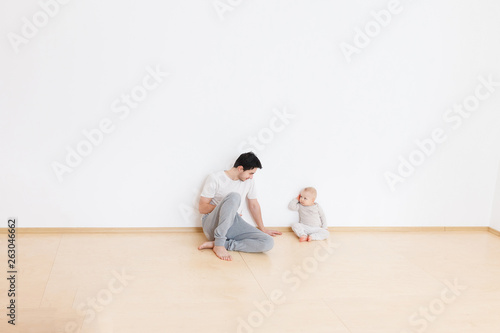 Paternal care of a little son. Little kid sits next to positive cute young dad talking and playing in cozy home atmosphere. White wall background