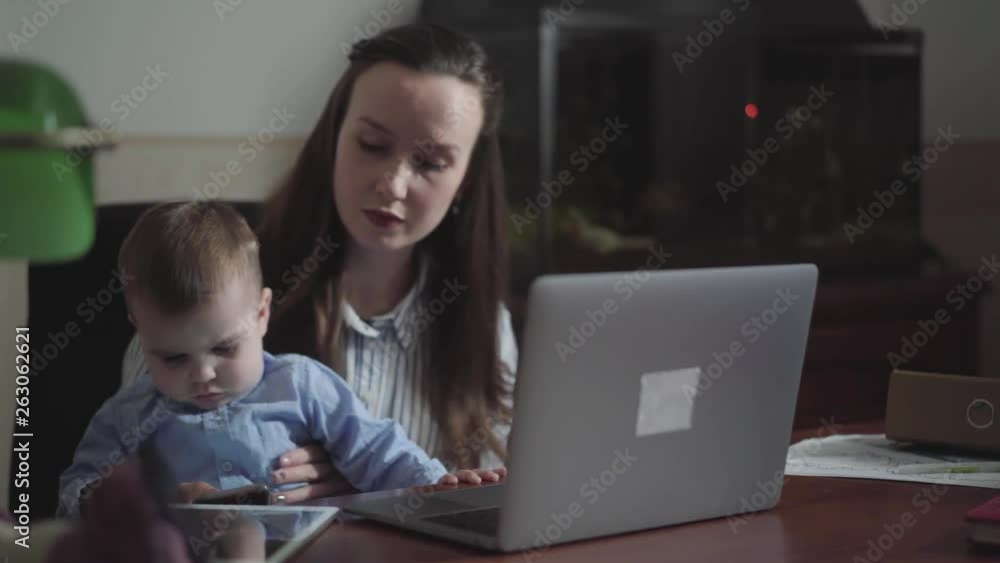Baby son sitting on mothers lap at the table touching cellphone in mom's hands. Busy young mother working at home. The child is accustomed to gadgets. Freelance concept. Camera moves left Baby son sitting on mothers lap at the table touching cellphone in mom's hands. Busy young mother working at home. The child is accustomed to gadgets. Freelance concept. Camera moves left