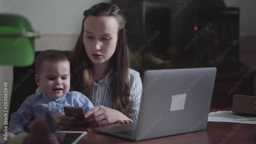 Portrait of mom and little son seated with a laptop at the table indoors. Busy young mother working at home. The child is accustomed to gadgets. Freelance concept. Portrait of mom and little son seated with a laptop at the table indoors. Busy young mother working at home. The child is accustomed to gadgets. Freelance concept.