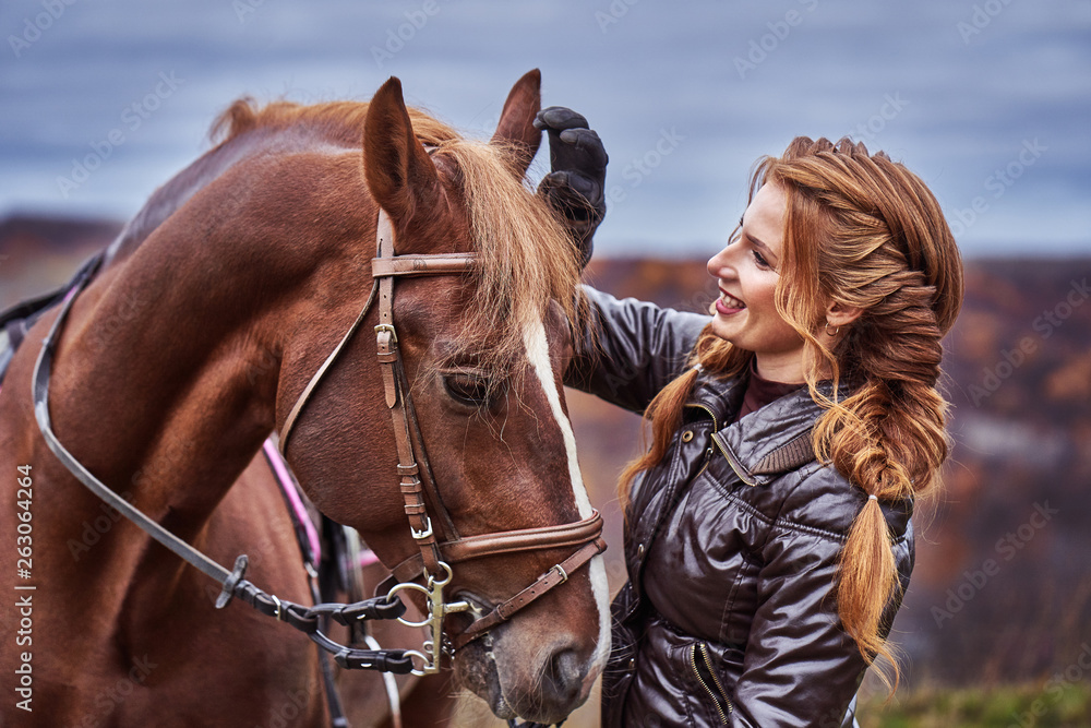 Obraz premium A young woman with brown curly hair caresses a brown horse. The horse caresses the woman. Woman on a walk with a horse on a cloudy autumn day.