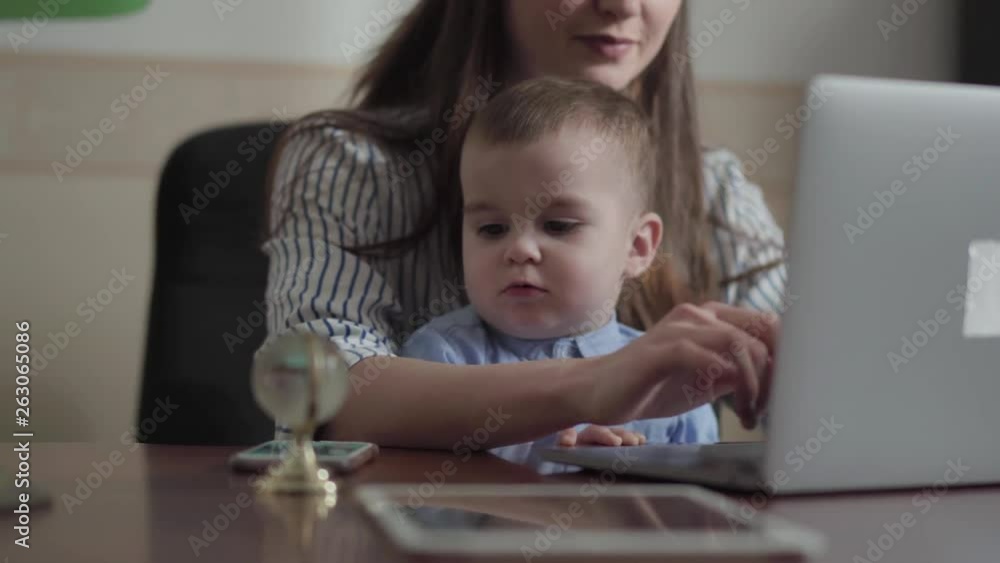 Portrait of pretty mother and little son seated with a laptop at the table indoors. Busy young mother working at home. The child is accustomed to gadgets. Freelance concept. Portrait of pretty mother and little son seated with a laptop at the table indoors. Busy young mother working at home. The child is accustomed to gadgets. Freelance concept.