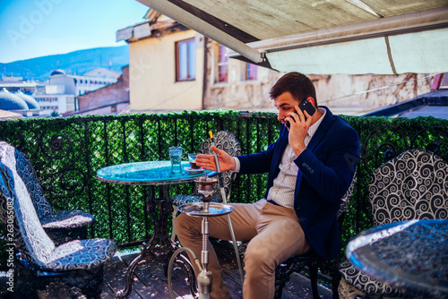 Young businessman wearing blazer and khaki smoking hookah at a coffee shop, while enjoying the amazing city landscape.