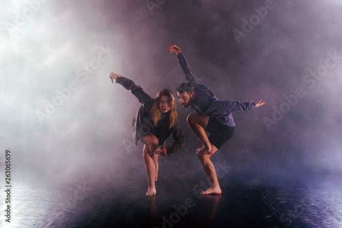 Two modern dancers stretching their shoeless feet high in the air surrounded by smoke on stage. © qunica.com