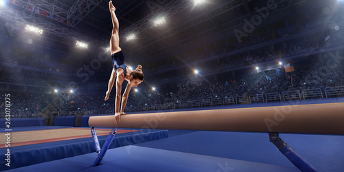 Female athlete doing a complicated exciting trick on gymnastics balance beam in a professional gym. Girl perform stunt in bright sports clothes
