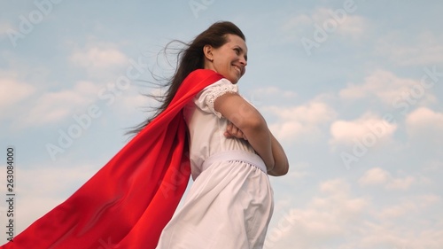 superhero girl standing on the field in a red cloak, cloak fluttering in the wind. close-up. girl dreams of becoming superhero. young girl in a red cape dream expression