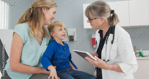 Senior woman pediatrician getting medical history from little boy patient and his mom. Child visiting doctor for regular checkup with his mother