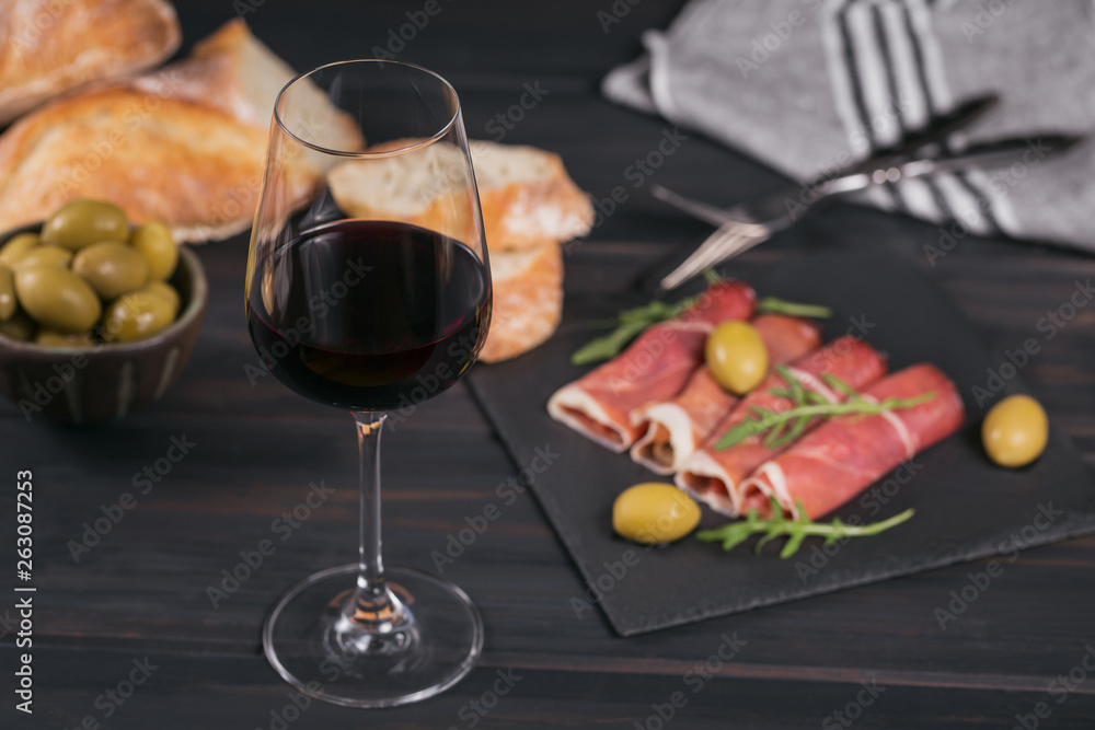 Glass of red wine with slices of cured ham or Spanish jamon serrano or Italian prosciutto crudo, bread, green olives and arugula