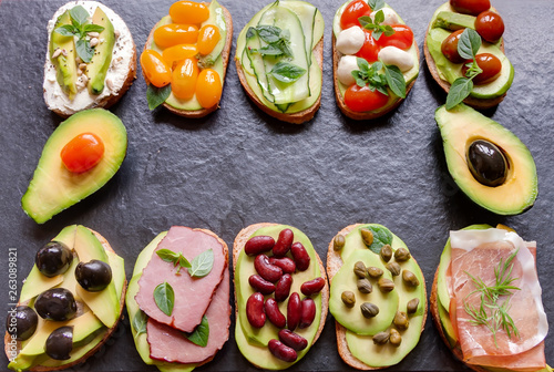Avocado sandwich top view with fresh sliced avocados, ham meat, mozzarella, cheese and vegetables on black background flat lay. Concept keto diet snack