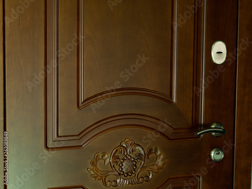 The entrance door to the house is brown with a pattern, handle and lock of gold color. The wooden door is of natural color.