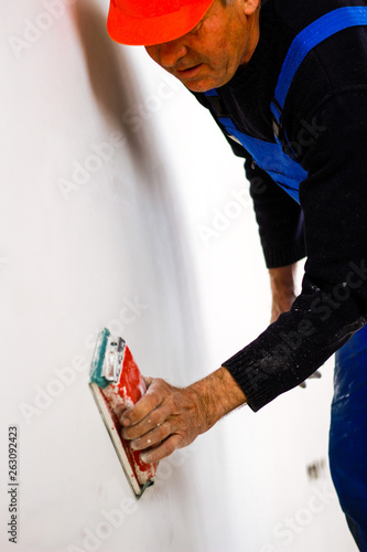 A builder in an orange building mask in his hands with a sandpaper holder. Blue form combining. Repair in the apartment.