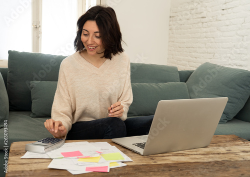 Happy young woman sitting on sofa surrounded by papers calculating expenses and paying bills