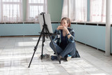 Girl artist paints with oil paints sitting on the marble floor. White canvas and easel stand on the floor of marble tiles in the room with turquoise and light green walls.