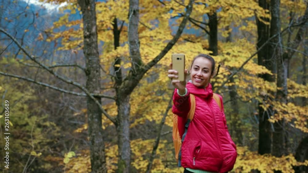 pretty woman hiker in red jacket with small backpack is standing on the yellow autumn woods background using phone and making selfie. pretty woman hiker in red jacket with small backpack is standing on the yellow autumn woods background using phone and making selfie.