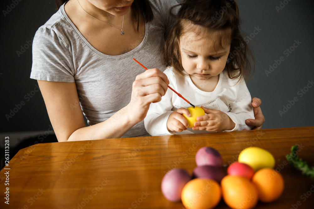 Fototapeta premium A young woman and her daughter are painting on an easter egg together. The girl is holding the egg while a woman is painting on it. Isolated on gray background.